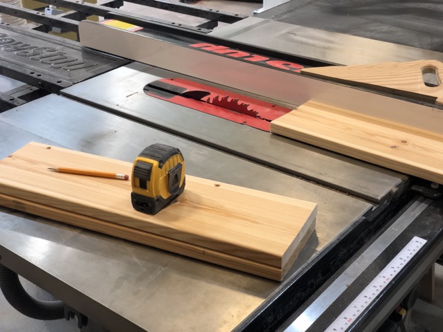An isometric angle of a table saw. The blade is slightly protruding and a small 20-inch board of wood sits in front of it in preparation for a cut. In the foreground out of the way of the blade are two identical boards stacked neatly with a pencil and tape measure resting on top.