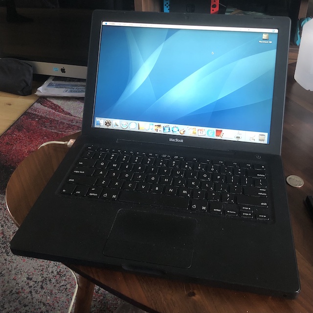 A matte black mackbook sitting on a walnut coffee table, booted to the default geometric blue wallpaper of MacOSX 10.4.
