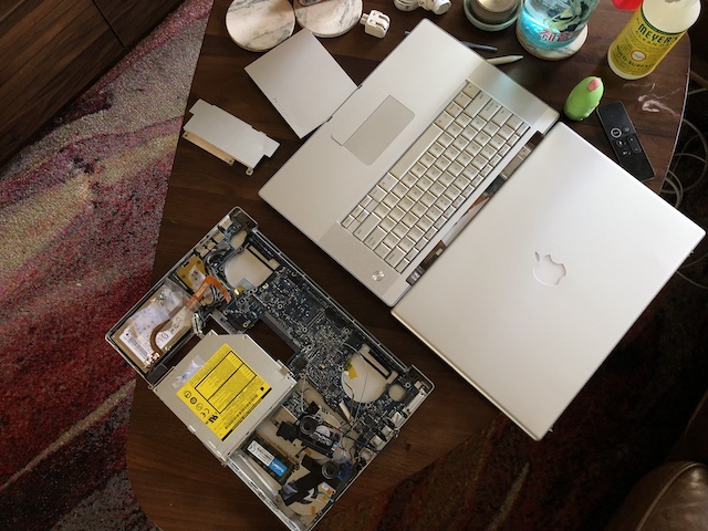 An old macbook pro disassembled in pieces on a coffee table; its guts exposed in the bottom case, with the keyboard and upper bezel sitting seperately next to it.