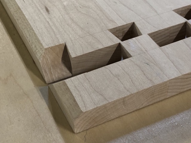 A closeup shot of two halves of a mitered dovetail joint. A longer board of wood that goes out of frame to the top left comes in at an angle to the center of the frame where a 45-degree corner is cut folowed by a few visible dove tails before they go out of frame to the right. Touching against the larger board is a very narrow smaller strip of wood where the receiving pins are cut laid flat on the table and touching the mating edge of the larger dovetails. The orientation of the two pieces of woods lets you visualize the smaller piece being rotated into the mating joint of the larger piece.