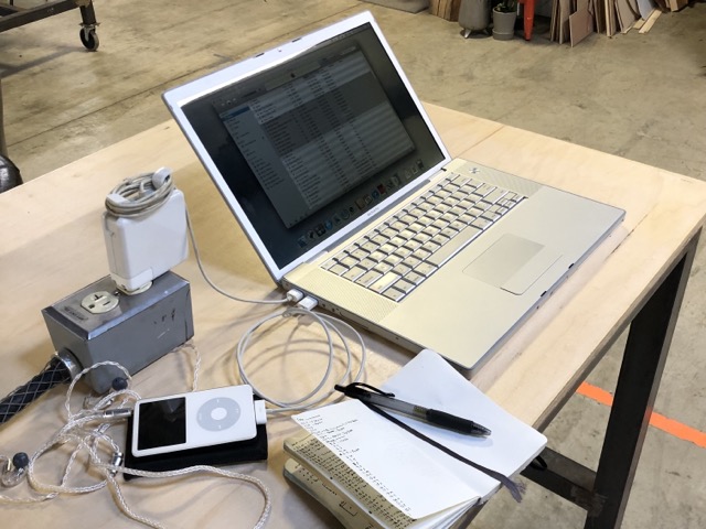 A 2006 MacBook Pro sitting on an industrial-style desk plugged directly into a charging brick on an outlet sitting next to it on the table. Connected via USB to the laptop is a crisp white 5th generation iPod video with an open pocket notebook sitting between them and hanging just out of the frame.