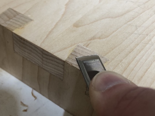 A close up shot of a sharp row of glued up dovetails cuts diagonally across the frame. In the center of the frame a finger pushes a sharp flush chisel through the top of one of the protruding pins bringing it in plane with the surface of the piece.