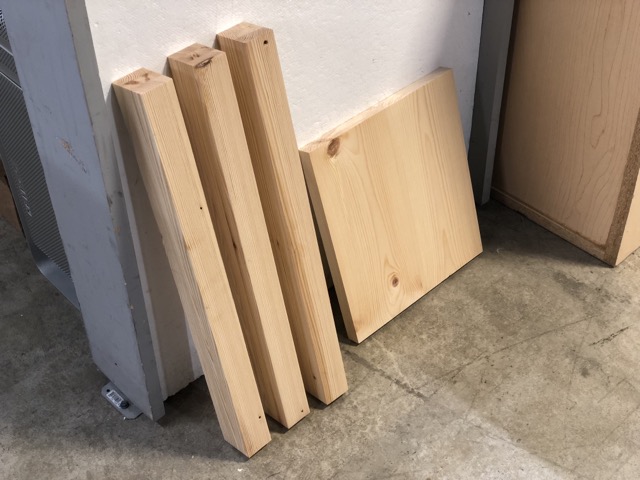 Three 20-inch laminated beams of pine rest against a white wall sitting next to a roughly 12-inch square by 1-inch thick panel of slightly nicer pine leaning at the same slight angle against the same wall.
