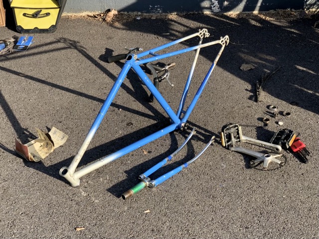 The frame and fork of a sky blue bicycle, with the headset and drop areas accented with silver. It is sitting on sunlit pavement, surrounded by sandpaper and many of the parts that were removed.
