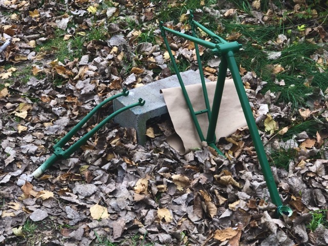 A bicycle frame and fork freshly painted british racing green, resting against a cinder block on top of long grass and fallen leaves.