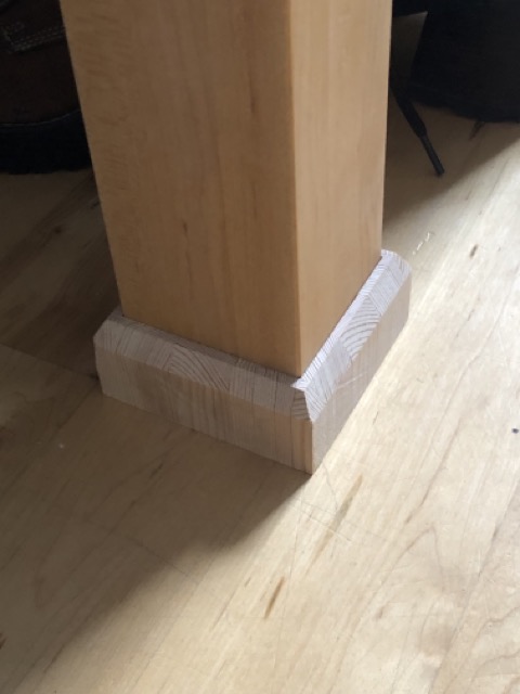 The rectangular leg of an oiled maple bed frame sitting inside of a chamfered, unfinished block of wood acting as a riser.